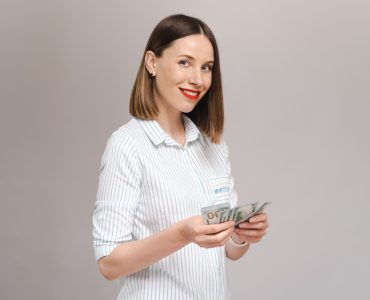 smiling young lady striped shirt holding money looking them gray wall looking front min